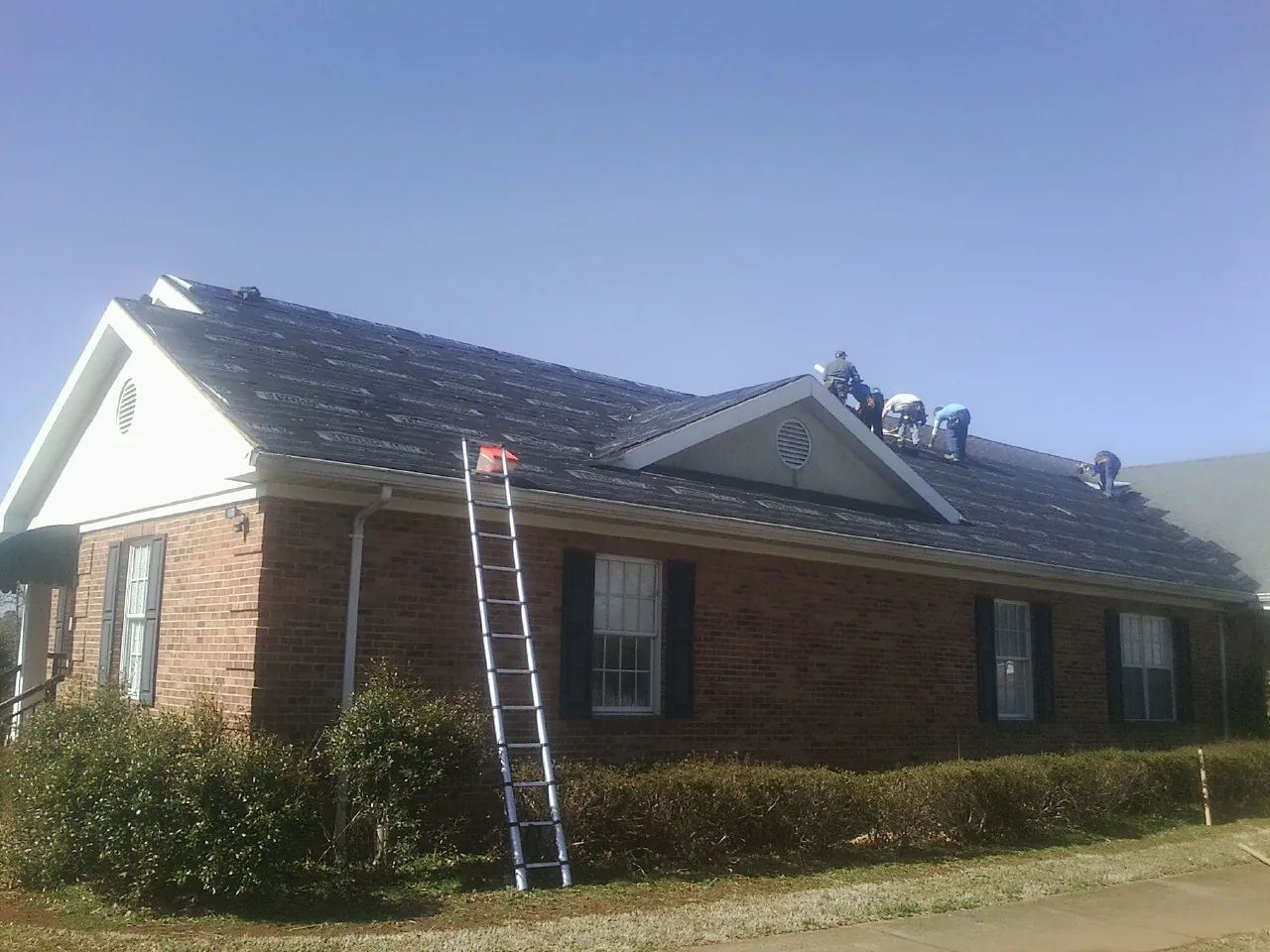 Crew tearing shingles off a brick ranch roof with a ladder set — Emergency Roof Repair work by our crew in Grosse Ile