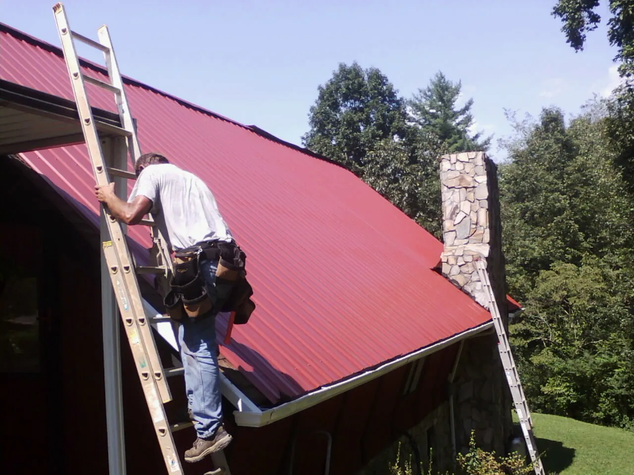Roofer on a ladder beside a red metal roof and stone chimney — Soffit & Fascia Repair by Roofing Brothers in Grosse Ile